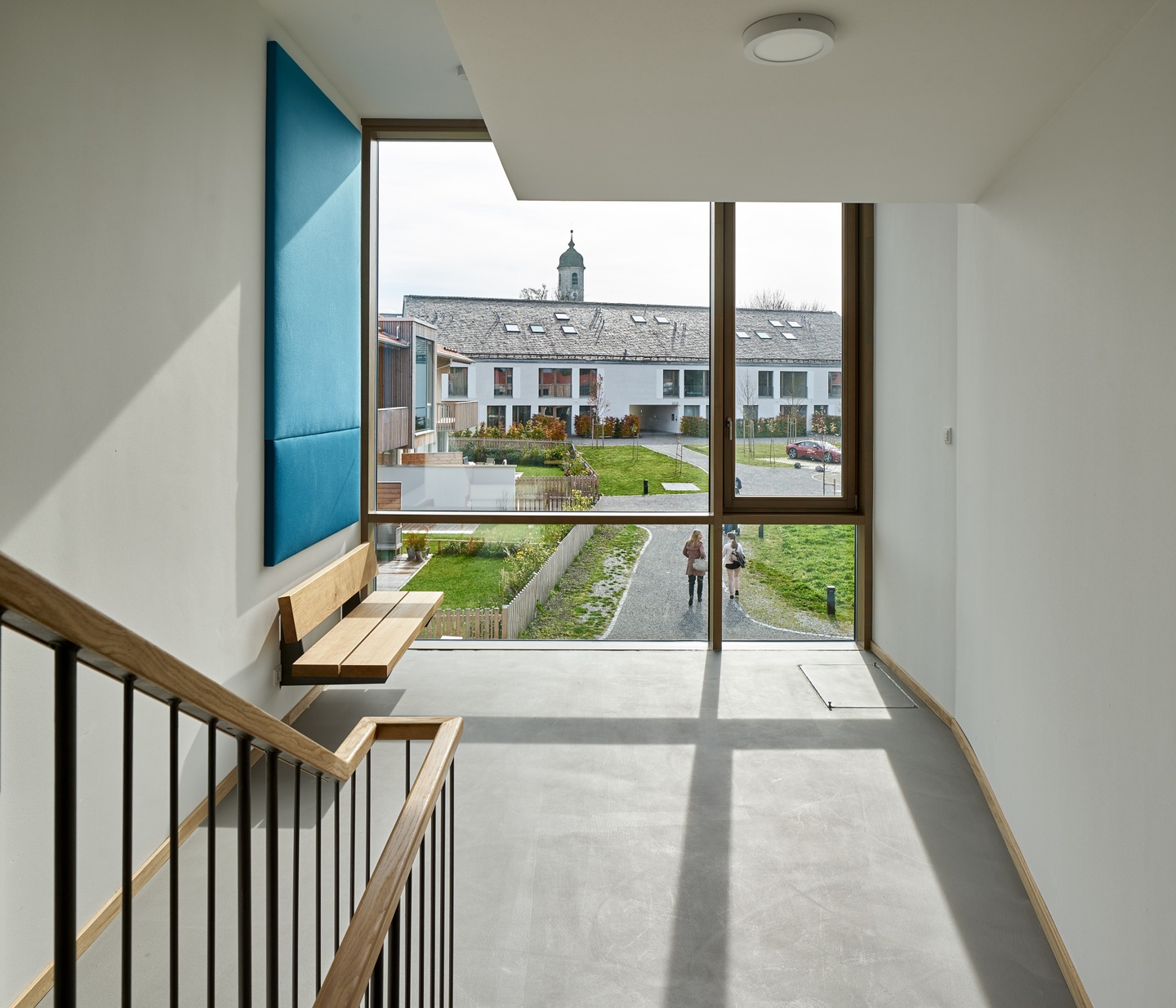 Interior of a house with white walls, concrete floor, wooden accents, and a window wall with mismatched panes looking out onto a walking path and other housing units.