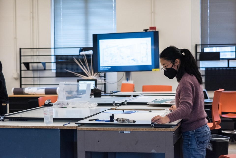 Person works at a long worktable with drawing implements on it. A tv screen is lit in the background displaying an architectural drawing.