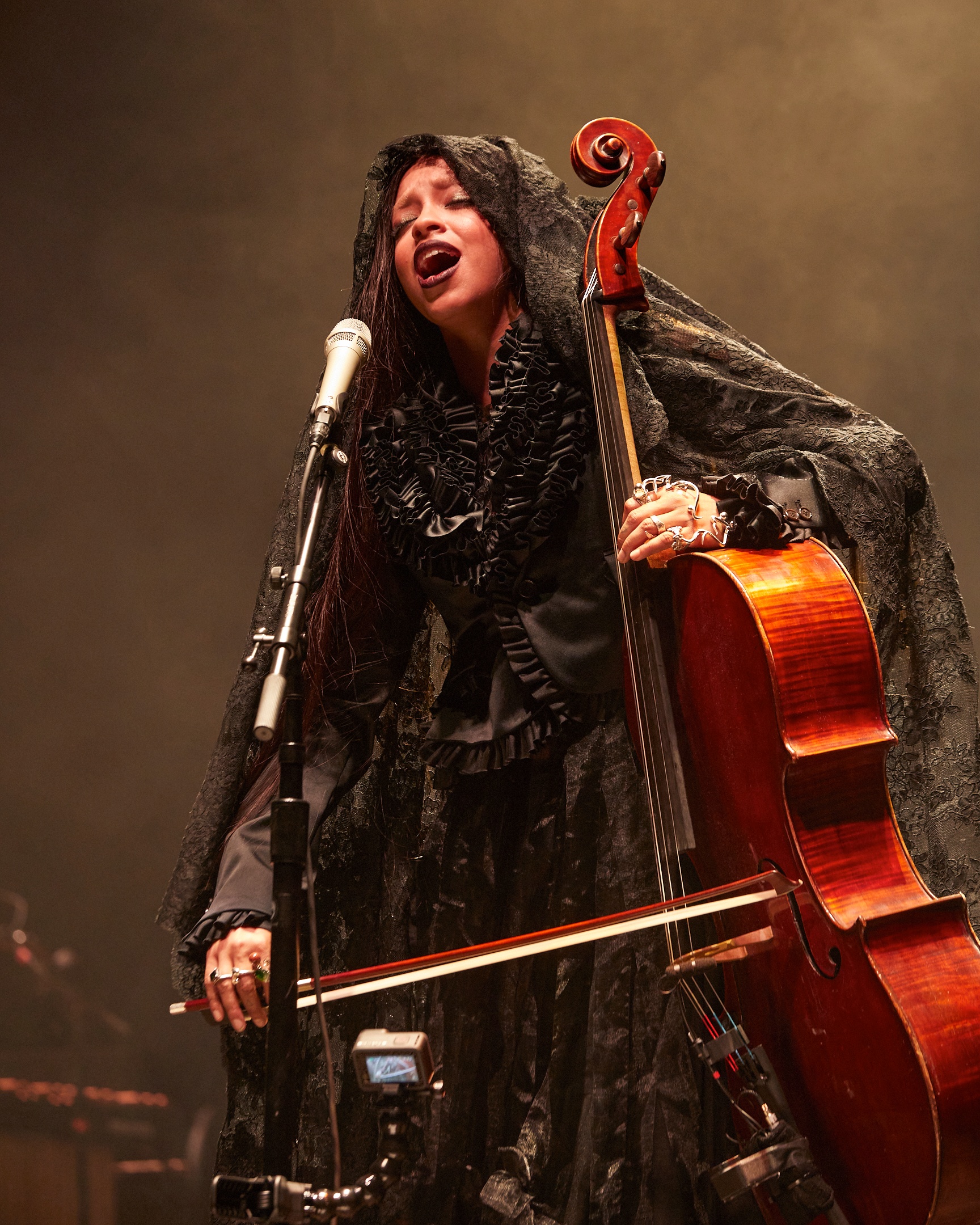 A Black person, the artist Kelsey Lu, performing on stage in front of a microphone that they sing into. They are wearing a black ruffled dress and lace scarf over their head, while holding a cello upright and playing it.