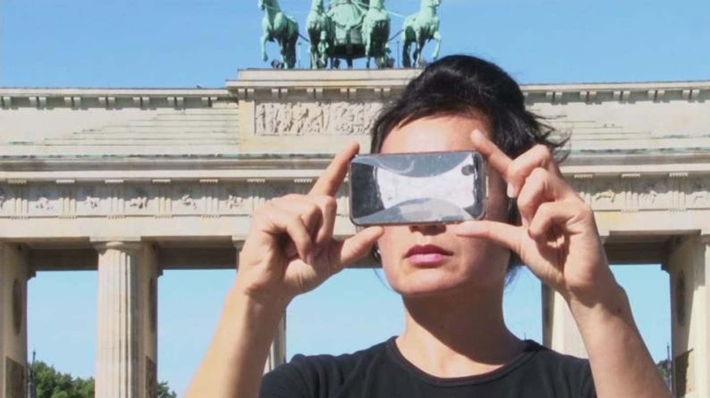 Video still of an individual taking a photo with their phone held horizontally; in the background is a blue sky and a monument-like structure with columns and a teal statue atop. 