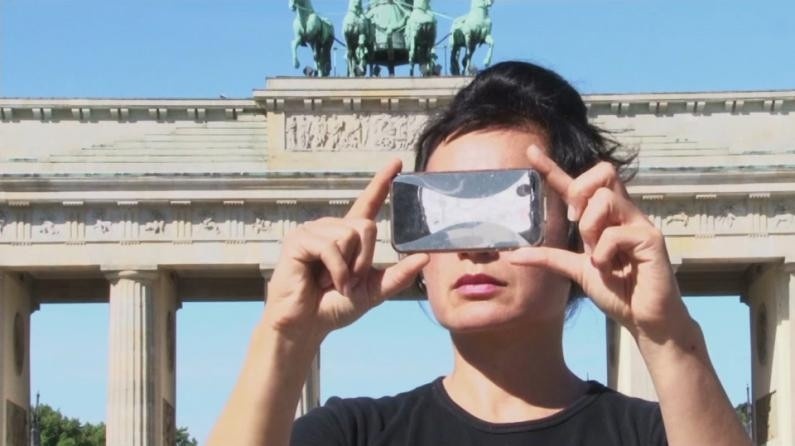 Video still of an individual taking a photo with their phone held horizontally; in the background is a blue sky and a monument-like structure with columns and a teal statue atop. 