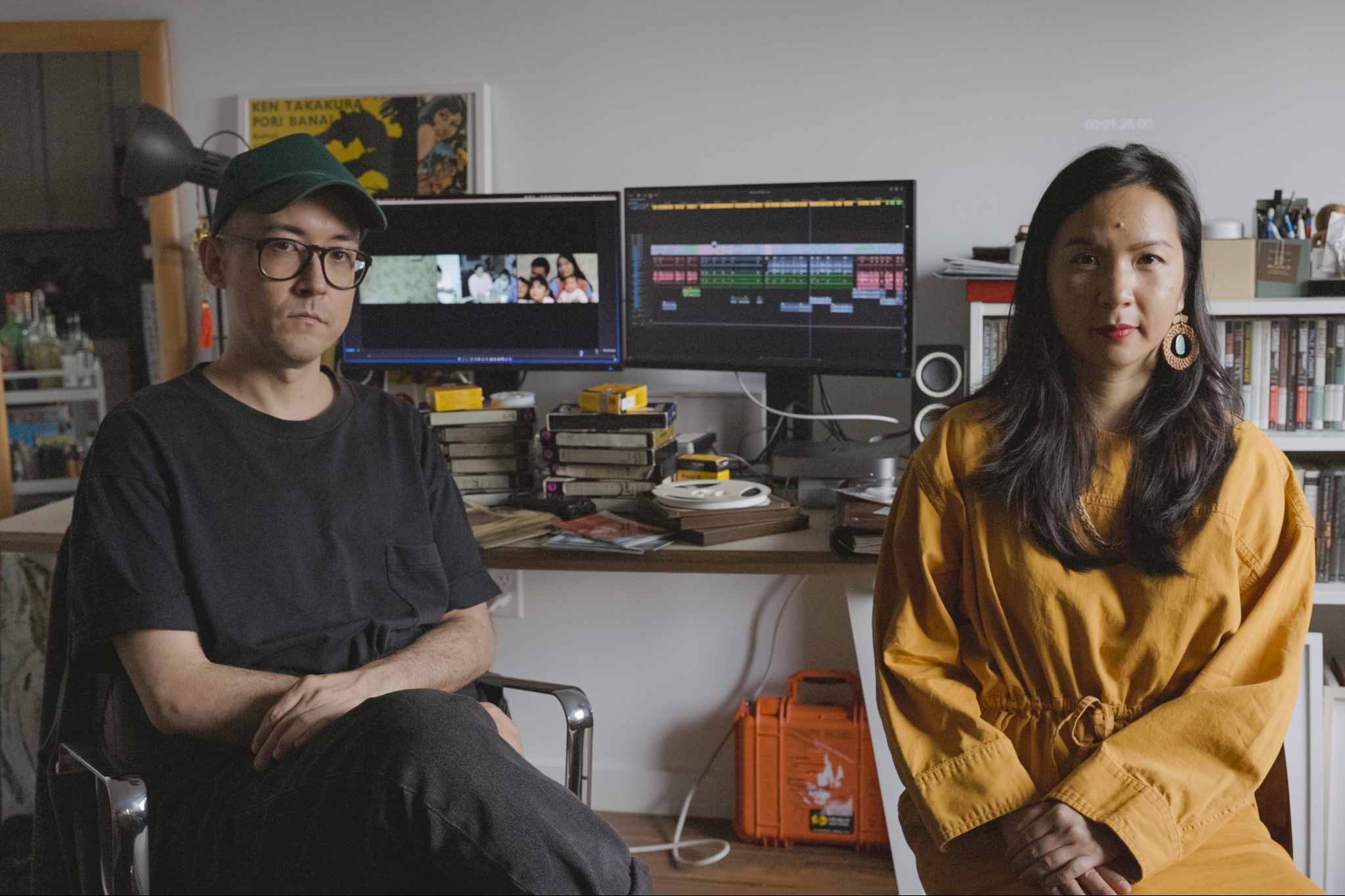 Artists Chritstopher Radcliff and Cathy Linh Che sit in front of a desk with two computer monitors. On one monitor is their film and on the other is the audio track for the film in an editing software program. On the desk are piled family photos and old VHS tapes from Che's family.
