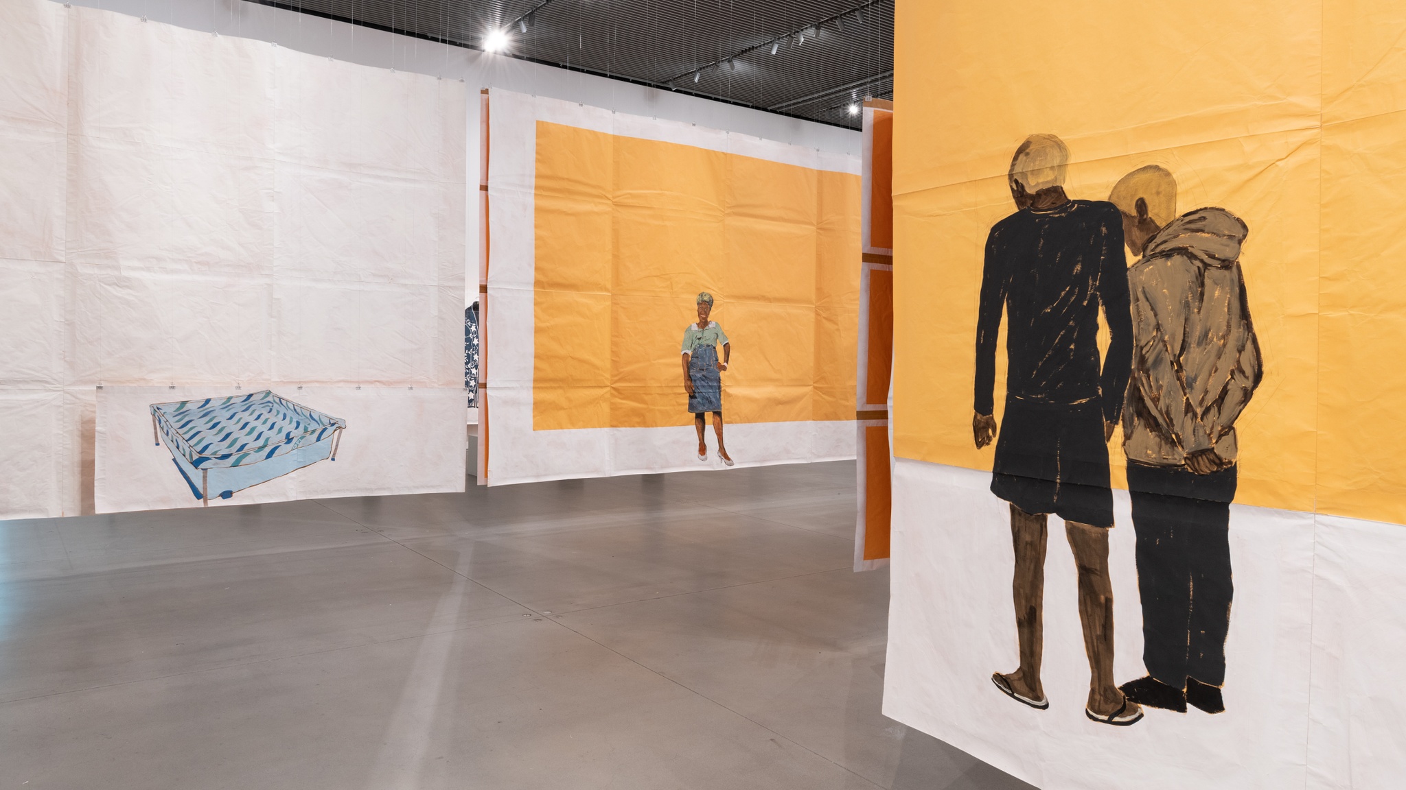 An art gallery with paintings on paper that hang from the ceiling to form passages. A corner formed by paintings reveals a painting of a Black woman posing against a yellow brown background. Another painting depicts a blue plastic pool against an all-white background. A final painting in the foreground shows two young Black men standing together with backs to the viewer. They both have blond hair.