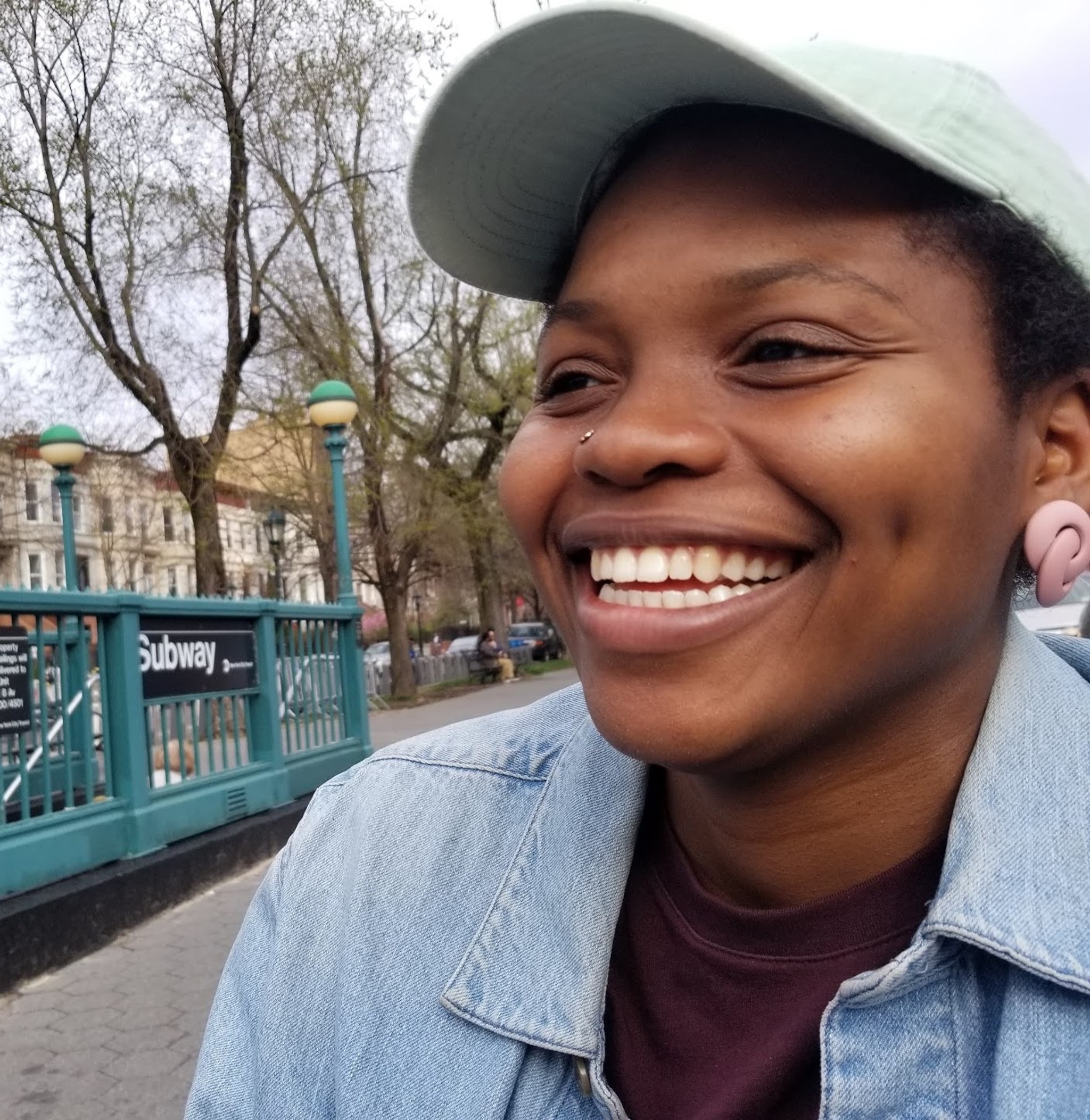 A close-up photo of a smiling Black woman, She has short hair under a pale green baseball cap and wears a blue collared shirt and pink earrings. In the background is the entrance to a New York subway station. 