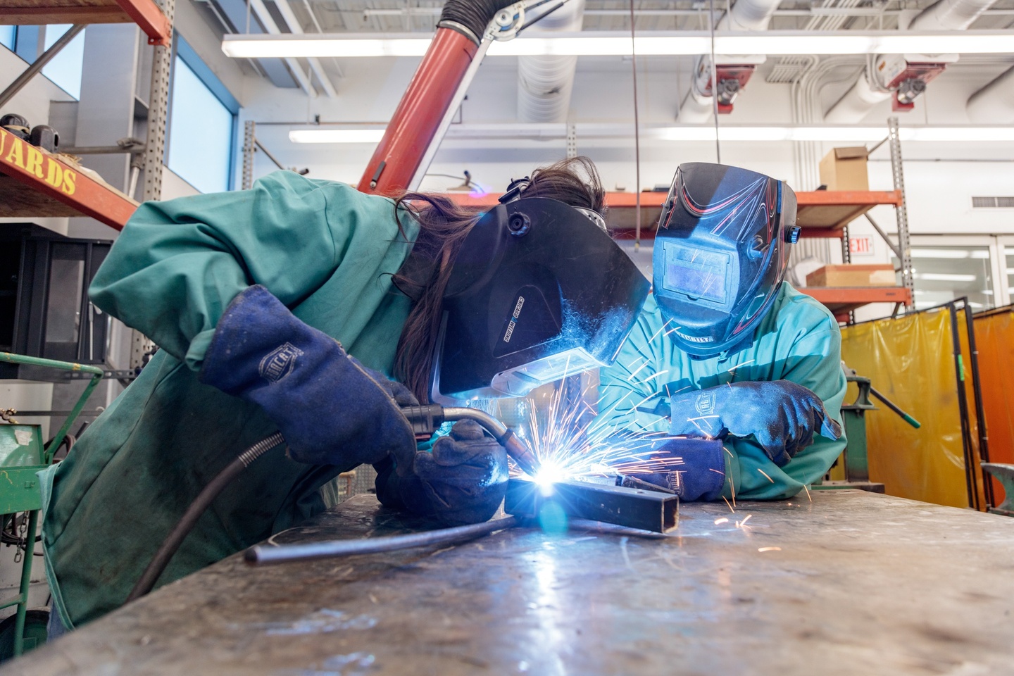 Two people in protective gear use a welder at a table in a high-ceilinged shop space.