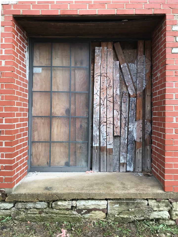 Entryway with a brick flat arch. Left half of the entryway has a glass door and the right half depicts drawings of faces on wood planks