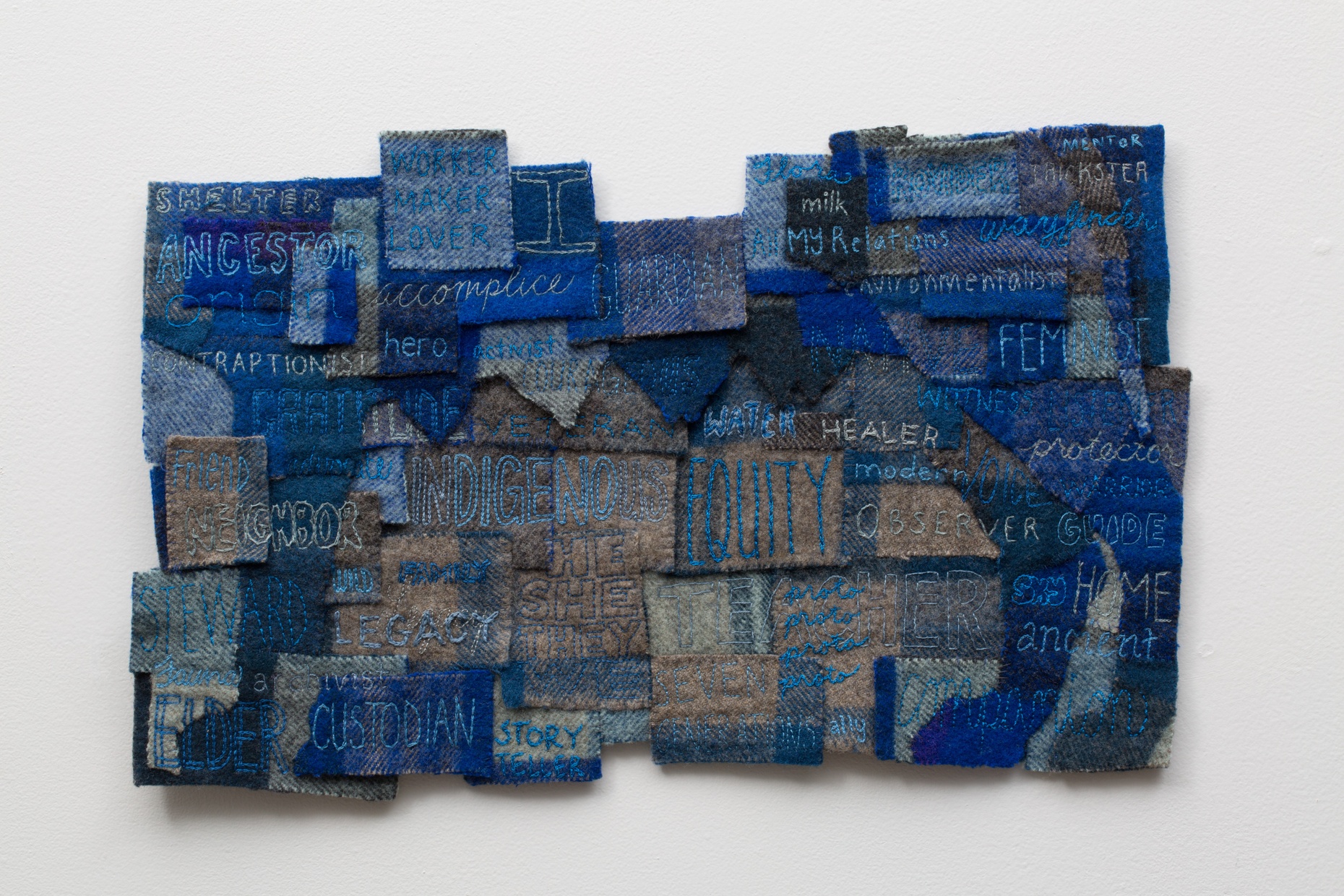 Squares of blue and brown fabric overlapping each other to make a large rectangle shape with words like "Indigenous," "equity," and "observe," embroidered on the squares.