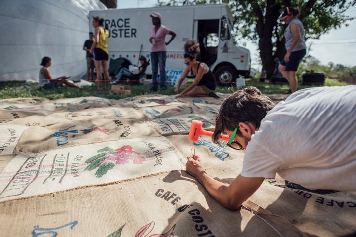 One person in the foreground hammers a stake into a coffee bag on the ground while other people work in the background.