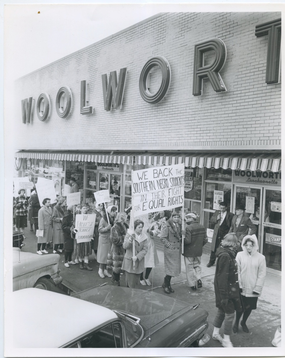 A black and white image depicts a line of light-skinned young women protesting in front of a Woolworth store. The protesters are wearing coats and holding signs and there are cars parked in front of the store.