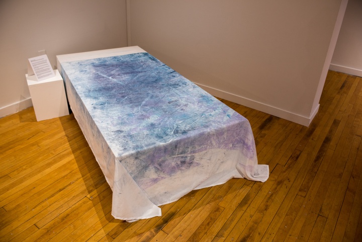 A white bedsheet is placed over a twin bed sized platform. The bedsheet is covered in blue and purple smudges, mostly towards the center of the sheet.