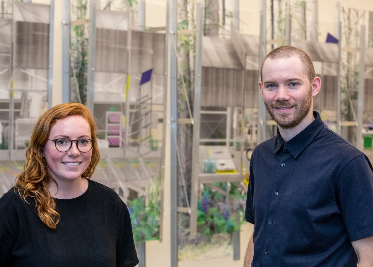 Two people stand in front of a paneled wall installation with architectural elements.