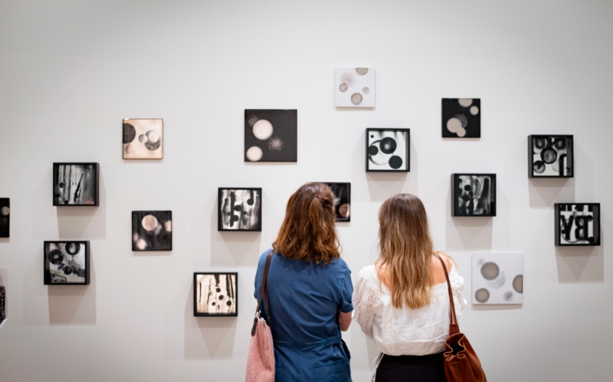 Two people with their backs to us face an installation of small, square, black-and-white compositions hanging on a wall