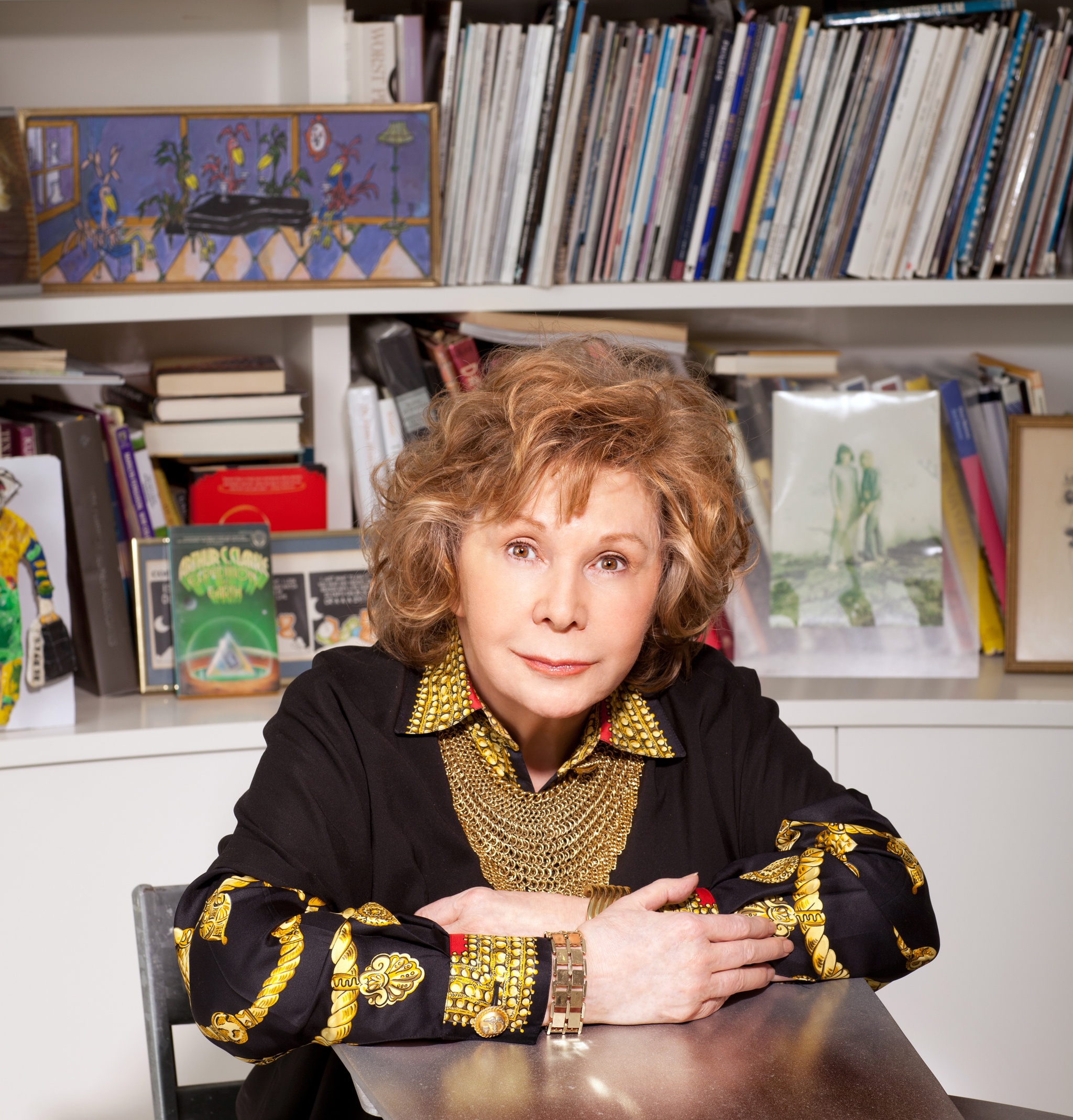 The writer Jane Wagner, a white woman with auburn hair, sitting at a table with hands crossed in front of her, with a shelf full of books and other objects behind her. 