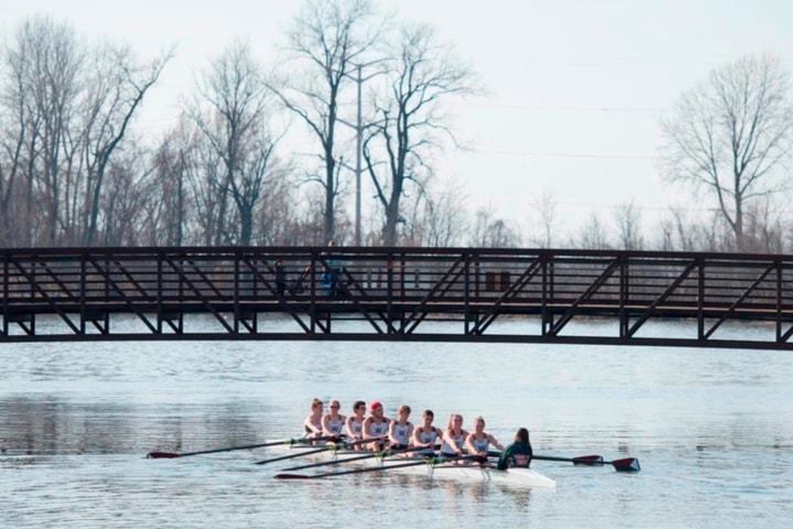A crew team rows under a bridge on a river in the winter