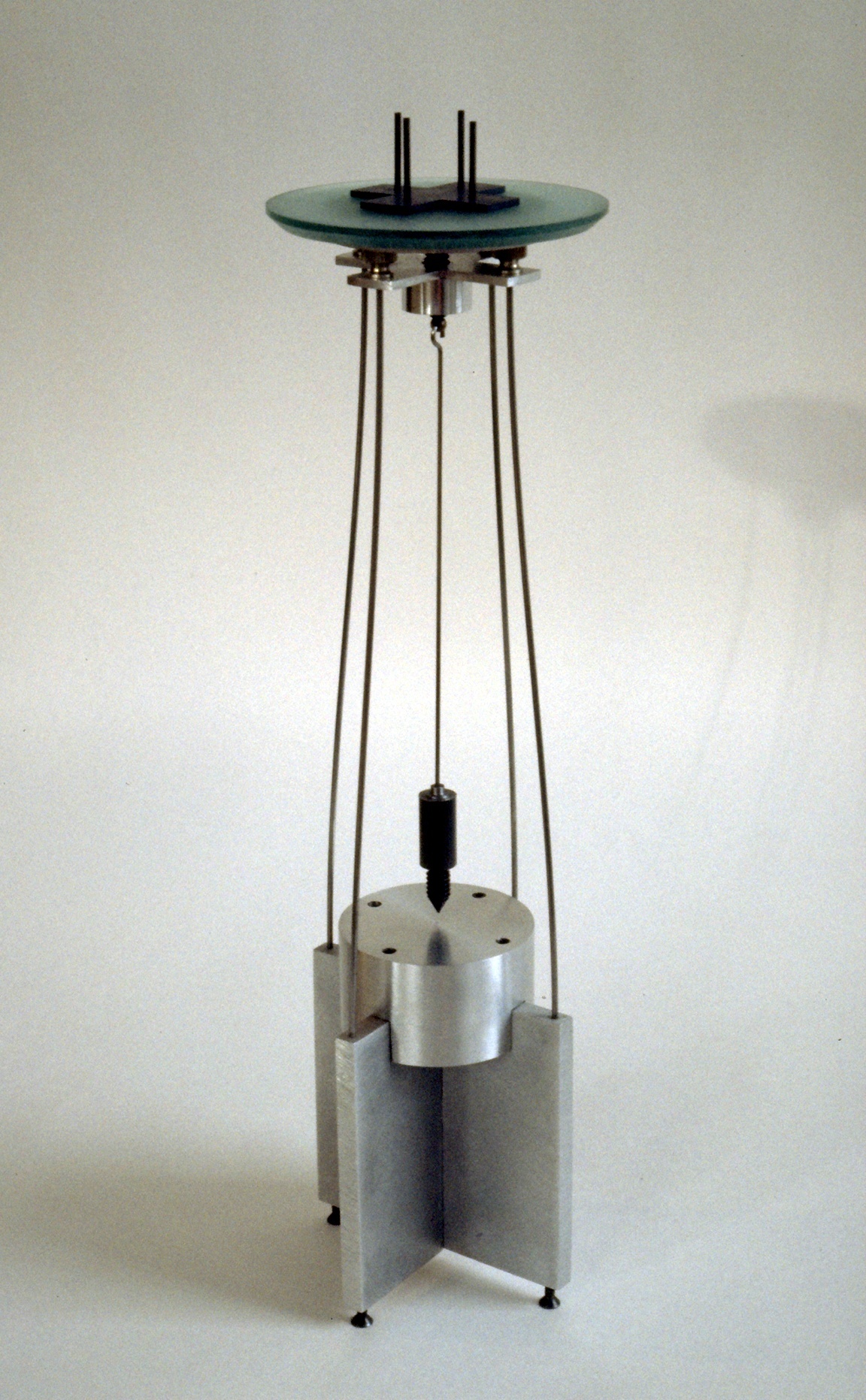 Small vertical sculpture with a weighted pendulum suspended from a glass disc over an aluminum base.