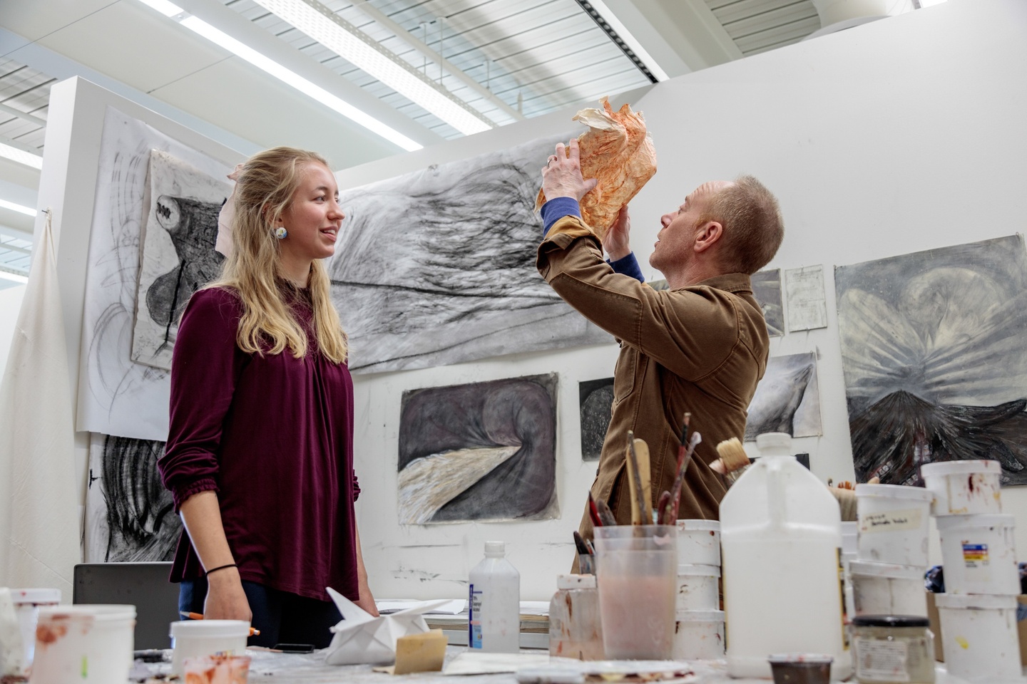 Professor and student in the student's brightly lit studio space; the professor is holding a sculptural object in the air and looking at it closely.