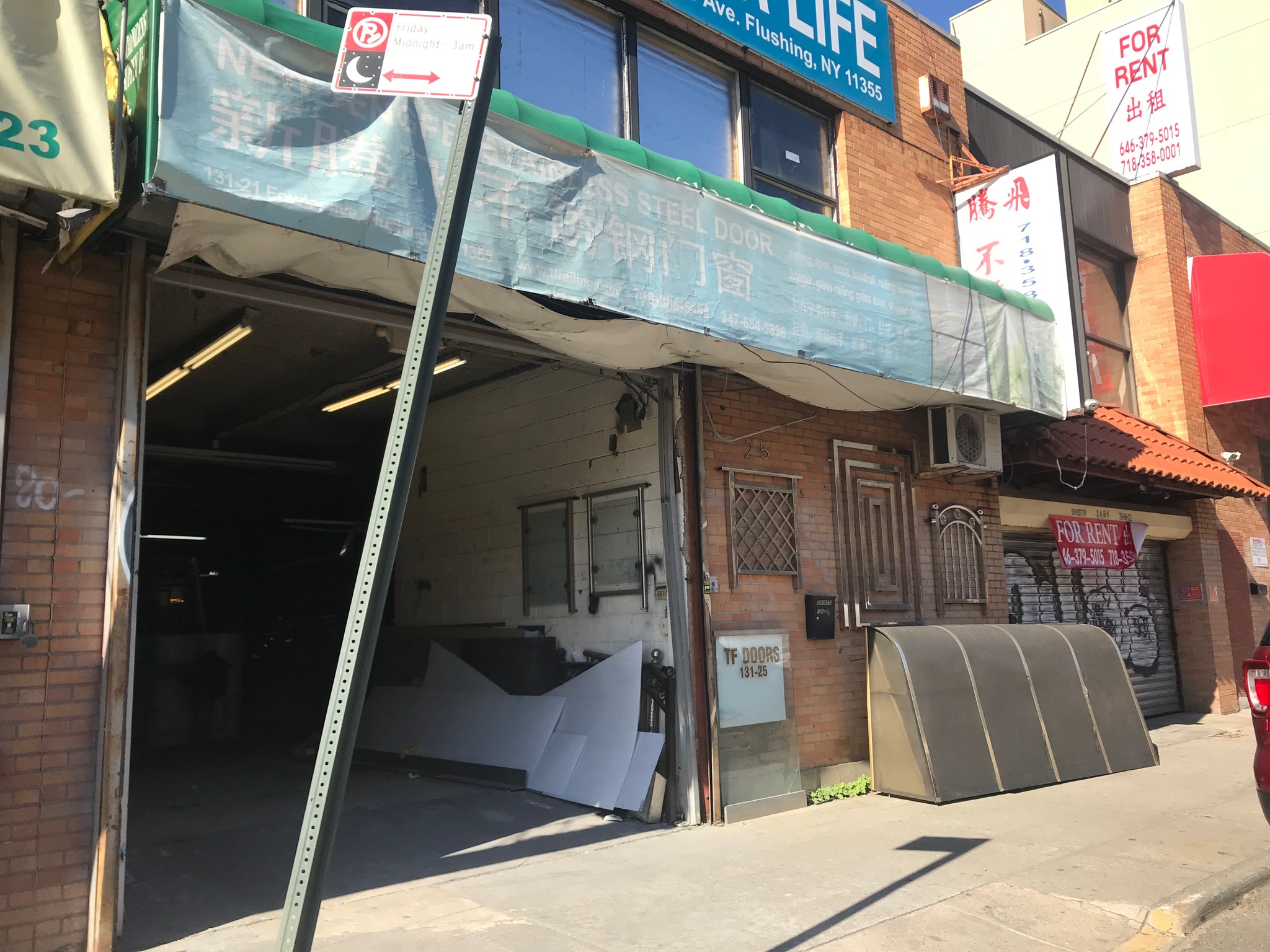 An open industrial garage door in the façade of a low, two-story row of brick buildings, with an assortment of commercial signs above and around the door. The space behind the door recedes into the building in shadow. The signs around the door are sun-faded and hard to read. Several read “For Rent” in red letters. 