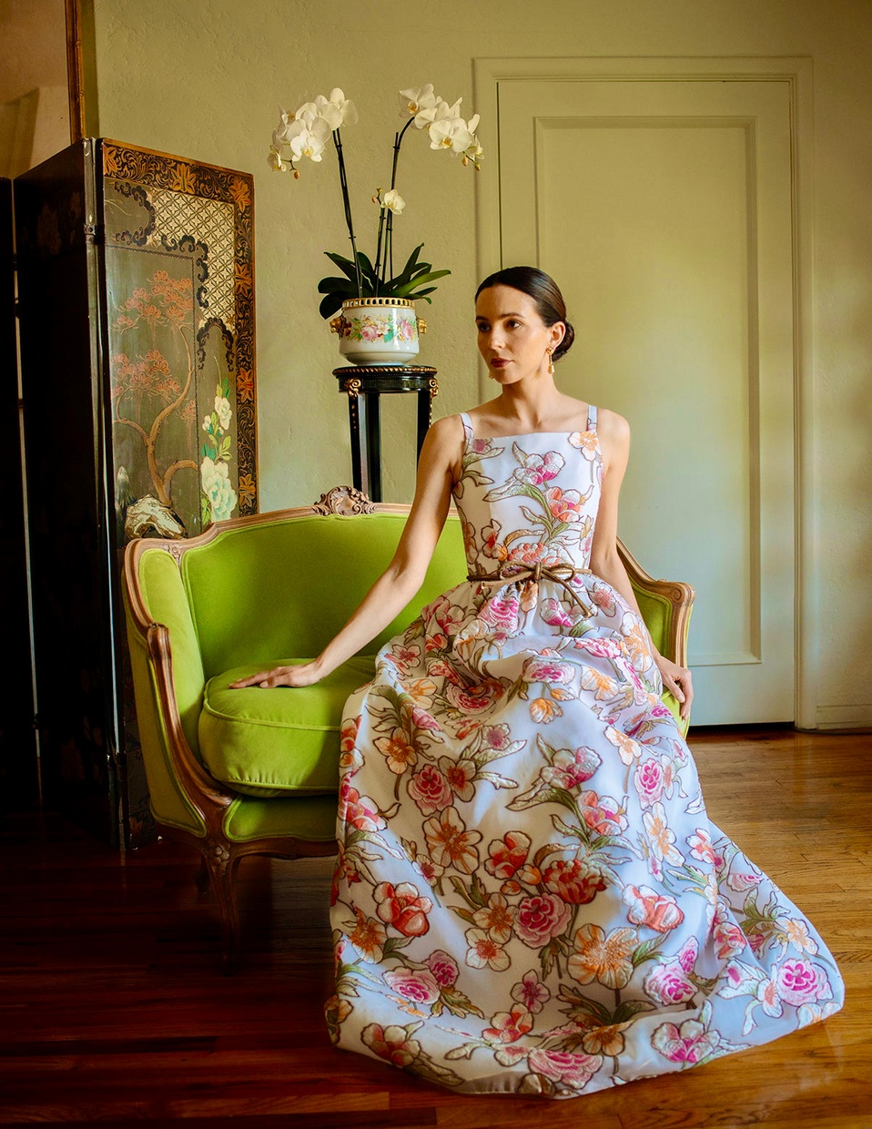 A seated woman looks off camera and wears a floor length, full-skirted, white dress with no sleeves that is embroidered with pink and orange flowers.