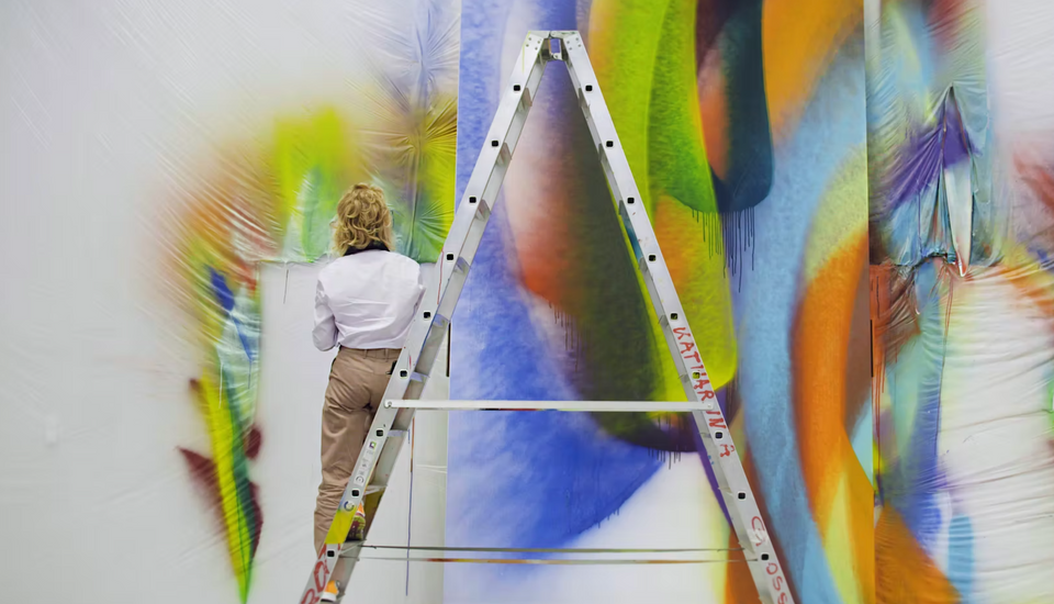 A person standing on a ladder facing a wall with colorful painted strokes