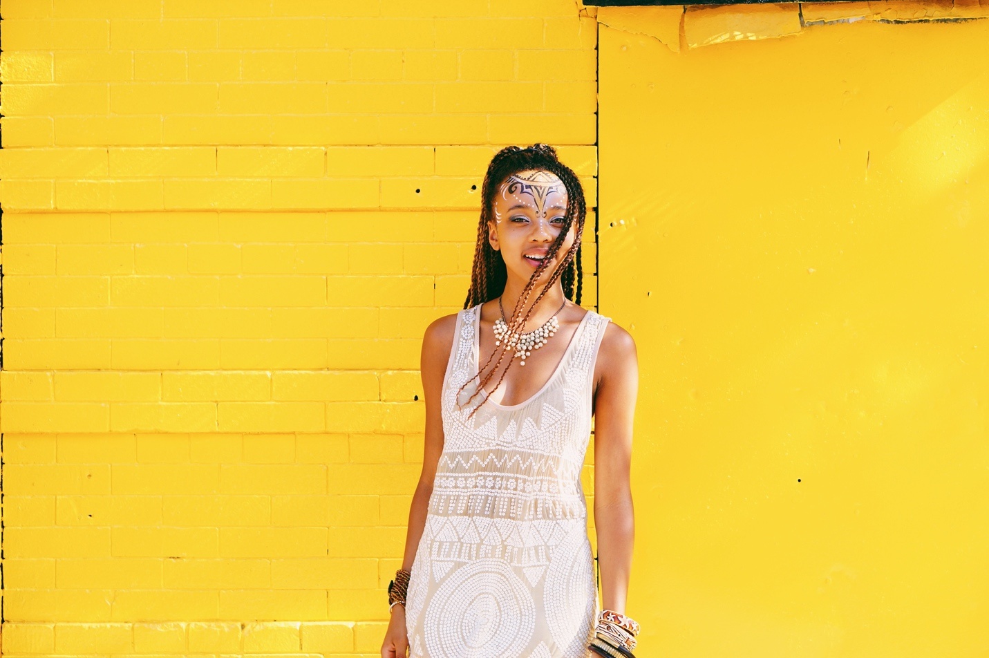 A photo of the artist Emily Waters standing against a bright yellow wall. They wear a white dress with arms at their sides as their braided hair fall across their smiling face.