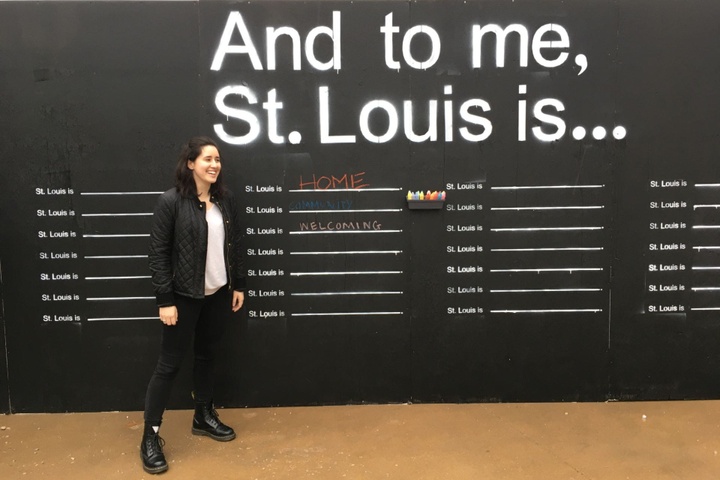 Eve Wallack stands in front of black chalk wall with large text: And to me, St. Louis is... with numerous lines for chalked responses.