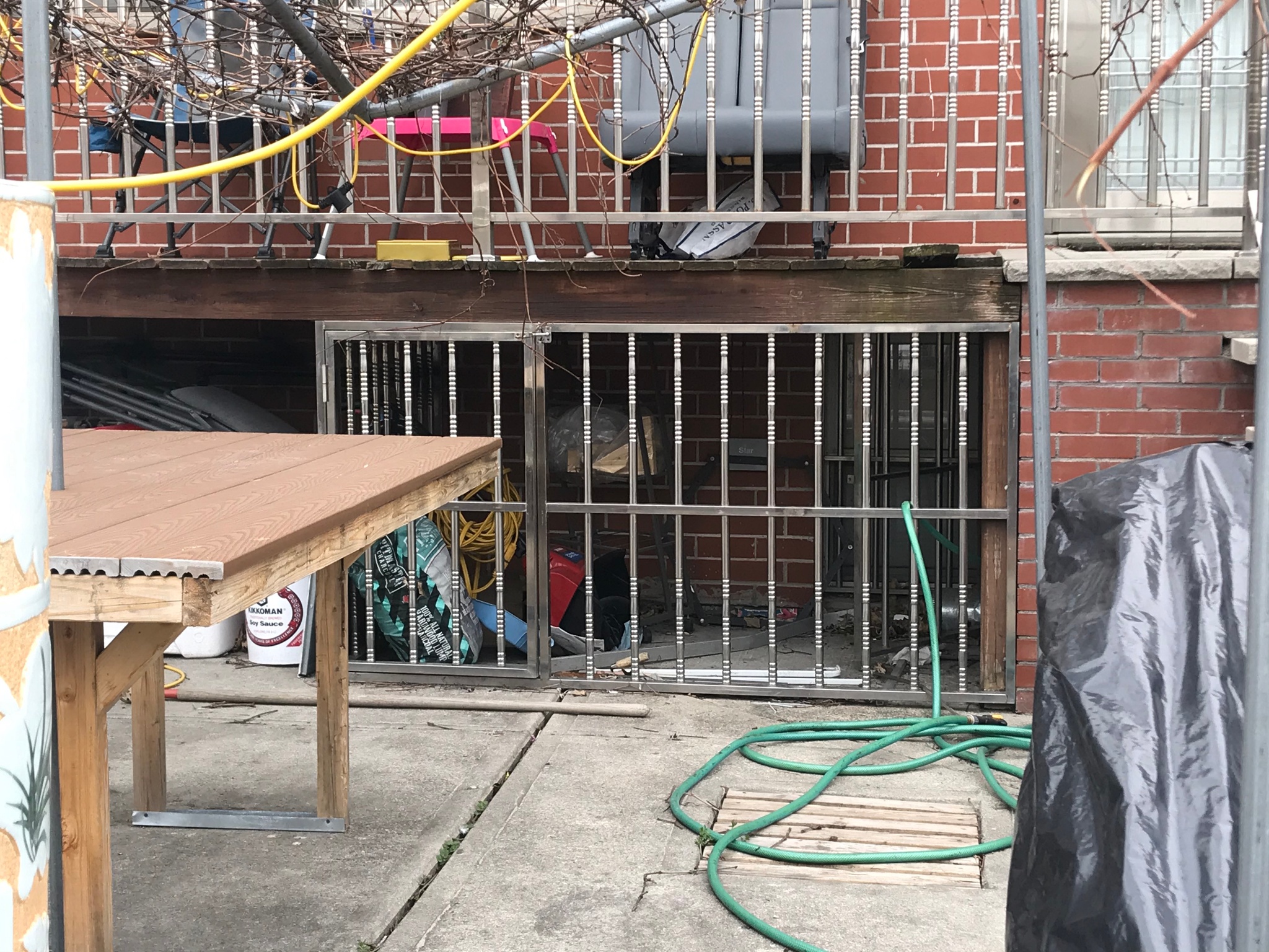 A crawl space beneath a narrow wooden porch attached to a brick building. The porch bisects the image horizontally and a stainless steel railing on the porch above matches a stainless steel gate that encloses the crawl space beneath it. A basement window is at the back of the crawl space and a green garden house snakes out of the gate.