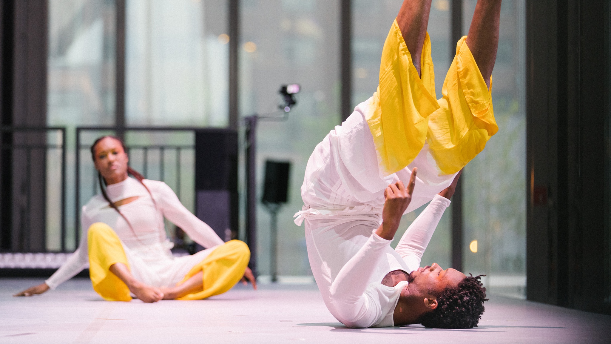 Two dancers on stage in white costumes with the legs dyed a bright yellow. In the background one dancer sits cross-legged on the ground. In the foreground the other dancer rolls backward on their back, lifting their legs high above their body.