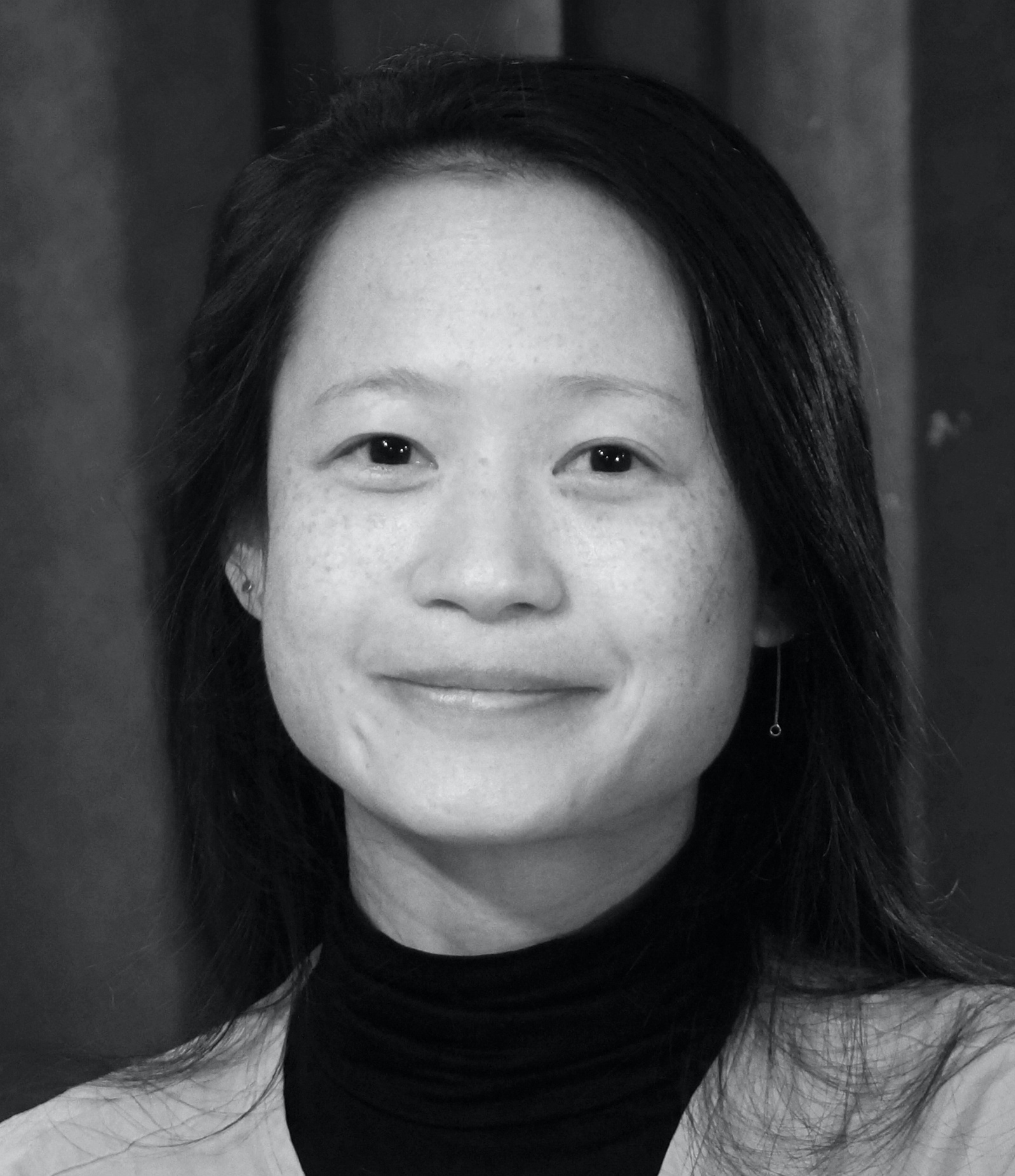 A black-and-white portrait of lighting designer Serena Wong, smiling with closed lips. She has straight black hair and wears a black turtle neck under a sweater lighter in color. 