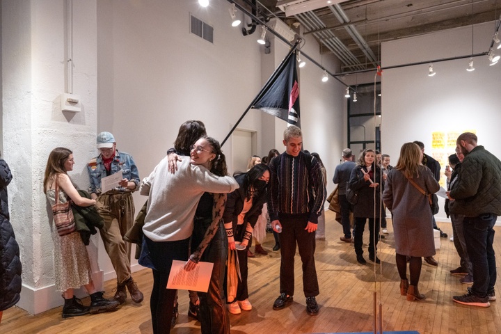 Two students embrace in a gallery space full of people. A handmade flag hangs above the crowd.