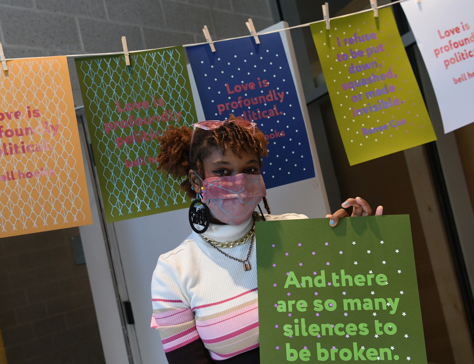 A masked, young, Black woman holds up a green poster printed with pink stars and lighter green text "And there are so many silences to be broken."