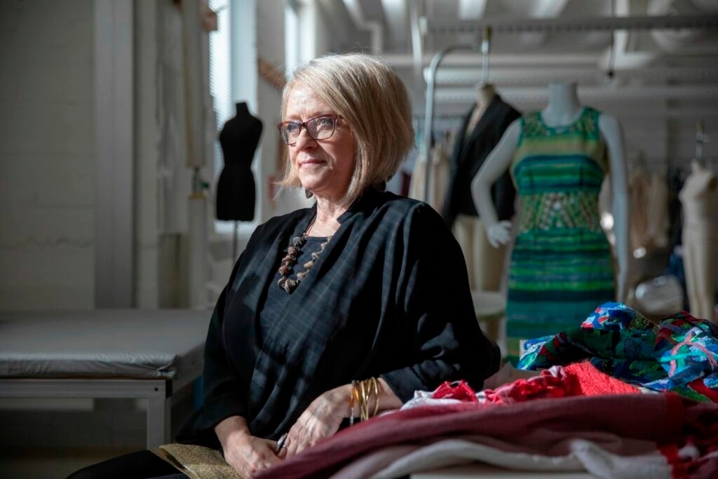 Portrait of Mary Ruppert-Stroescu in the fashion design studio. Mary sits to the right of a bank of large windows, her face sunlit. Her arm is propped on a table piled with various fabric samples. In the background are several mannequins, some of which are dressed in RECLEM garments.