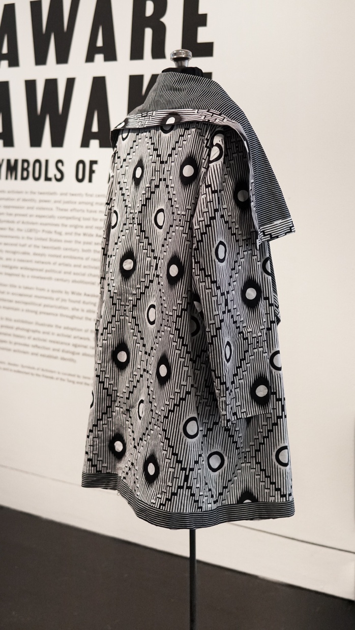 A long, black and white cape with sleeves and a large, fold-over collar; the fabric has thin, vertical stripes and evenly-spaced polka-dots centered within diamond shapes.