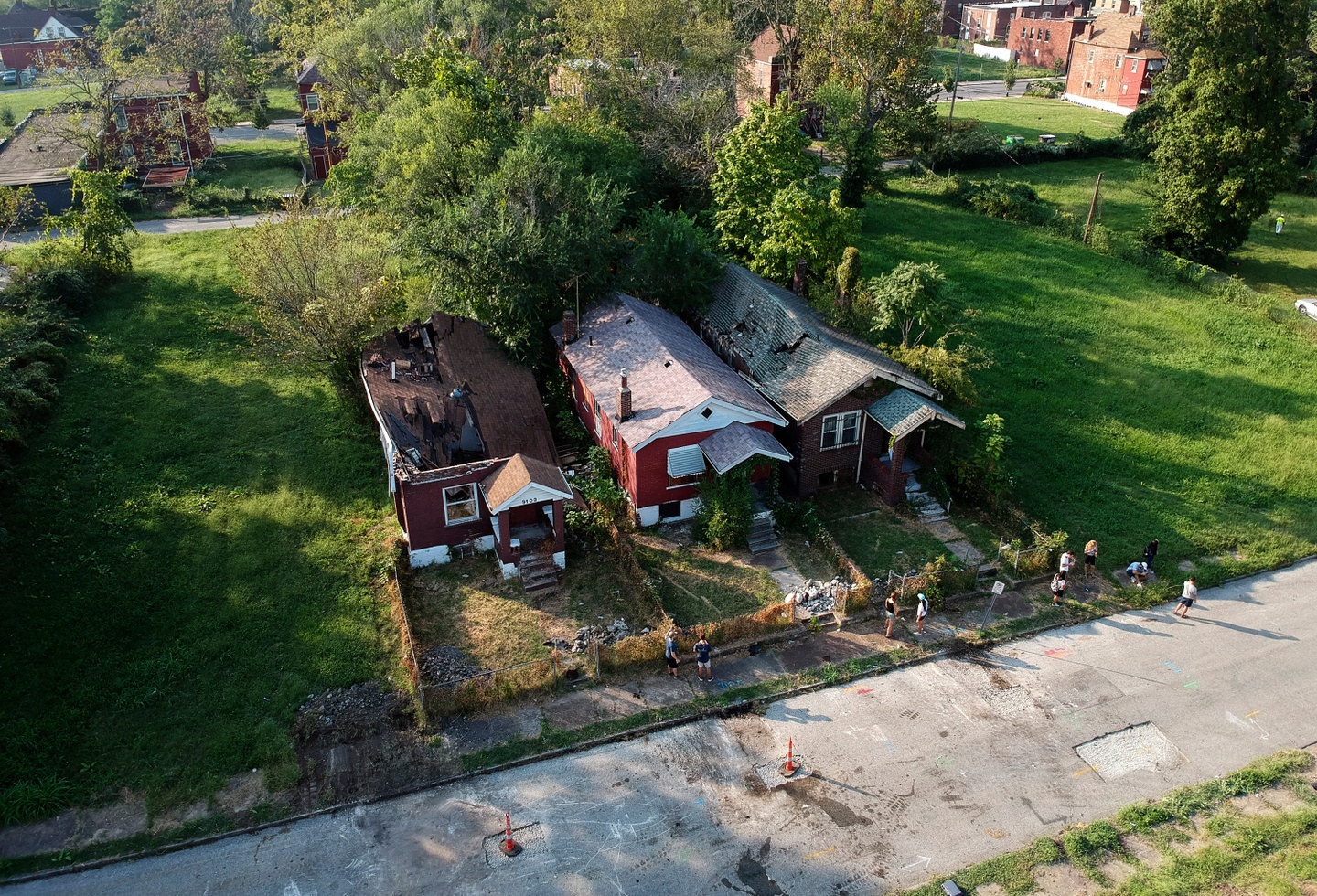 Drone photo of three brick houses side by side on a block, empty lots on either side, all with obvious damage to their roofs and structures and heavy overgrowth in the backyards.
