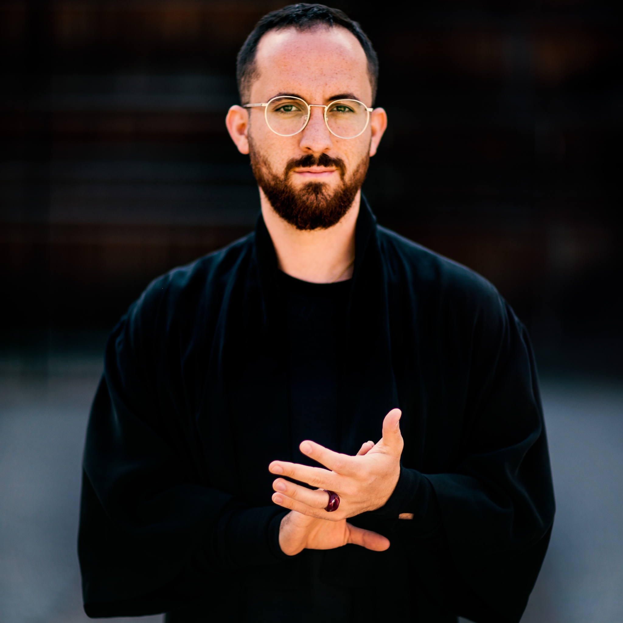 A white man stands facing us with hands held out in front of his chest. He wears dark clothes and wears glasses. 