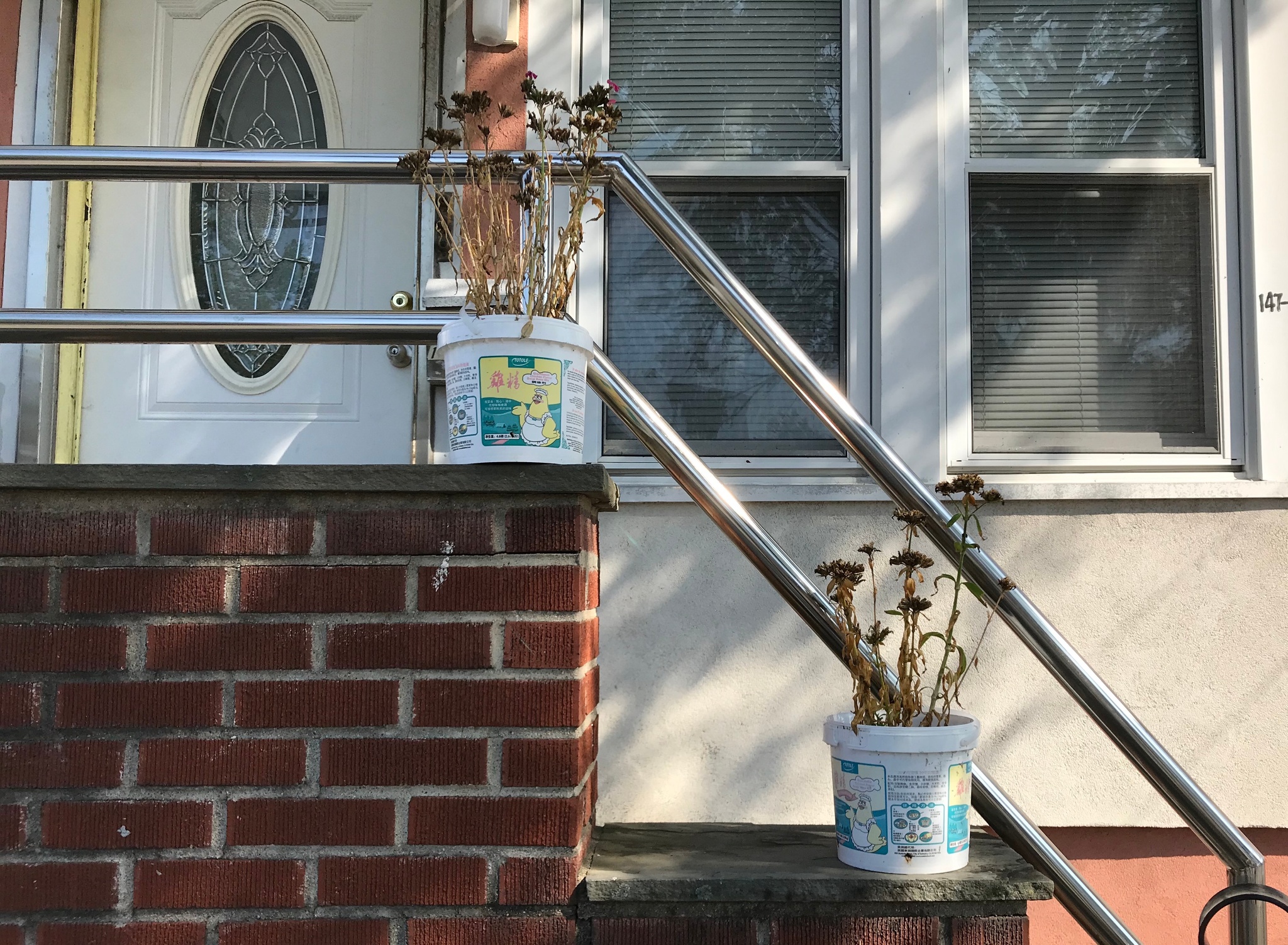 A front porch on a brick house with a stainless steel banister. Dead flowers sit in plastic buckets placed on the landings of two stairs. 
