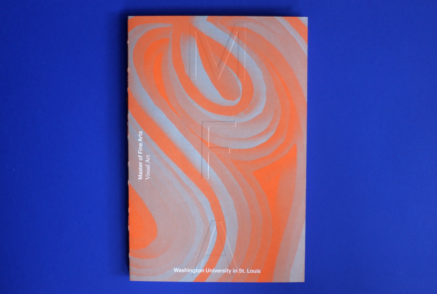 Orange and silver book photographed on a deep ultramarine background. The cover features an abstract, curved pattern with the letters "MFA" embossed in the center. Smaller text, in white, reads "Master of Fine Arts" — "Visual Arts" — "Washington University in St. Louis".