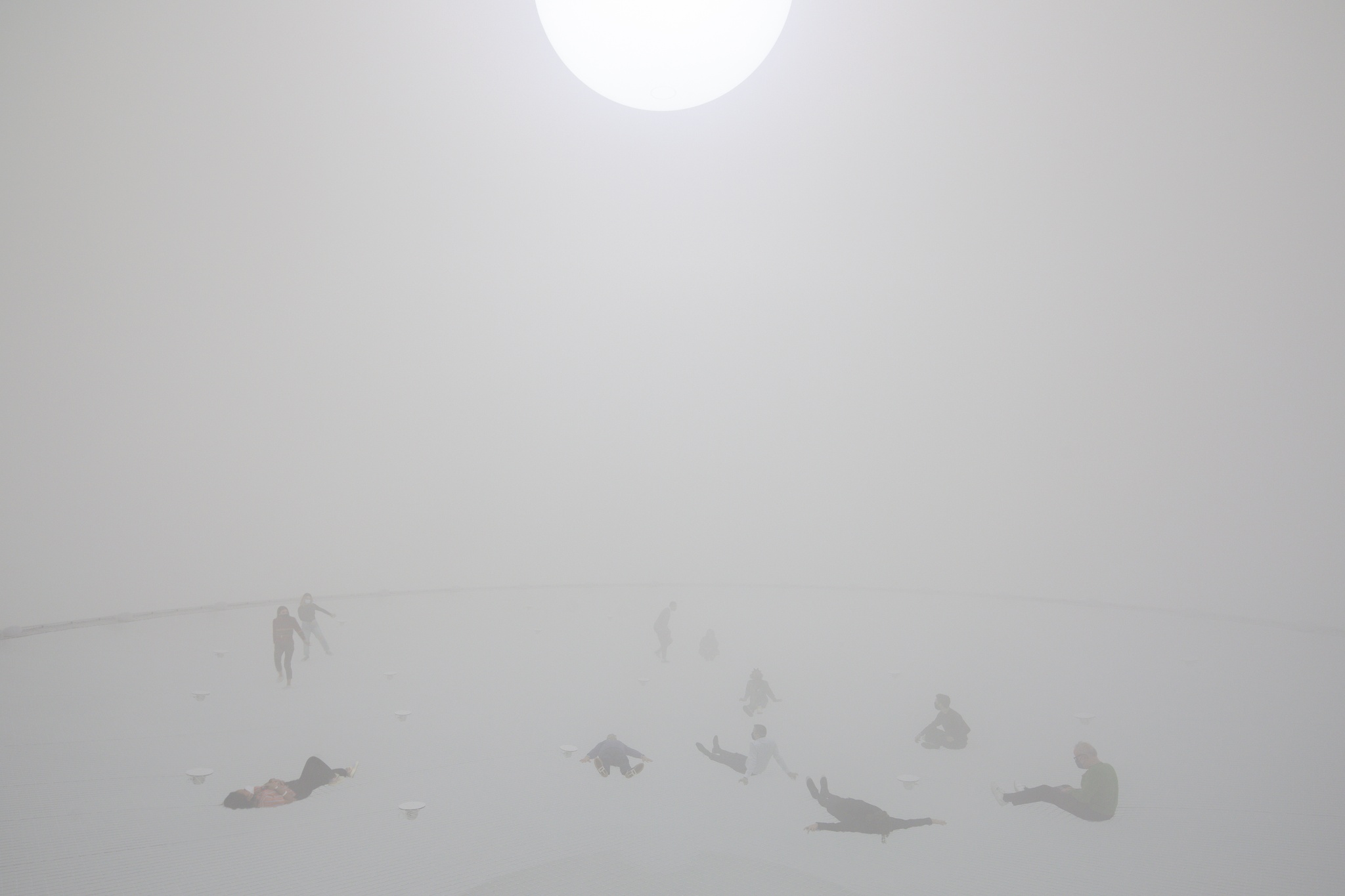 People sitting suspended on a net in a large spherical space that is illuminated by a large spherical white light above and filled with a foggy haze.