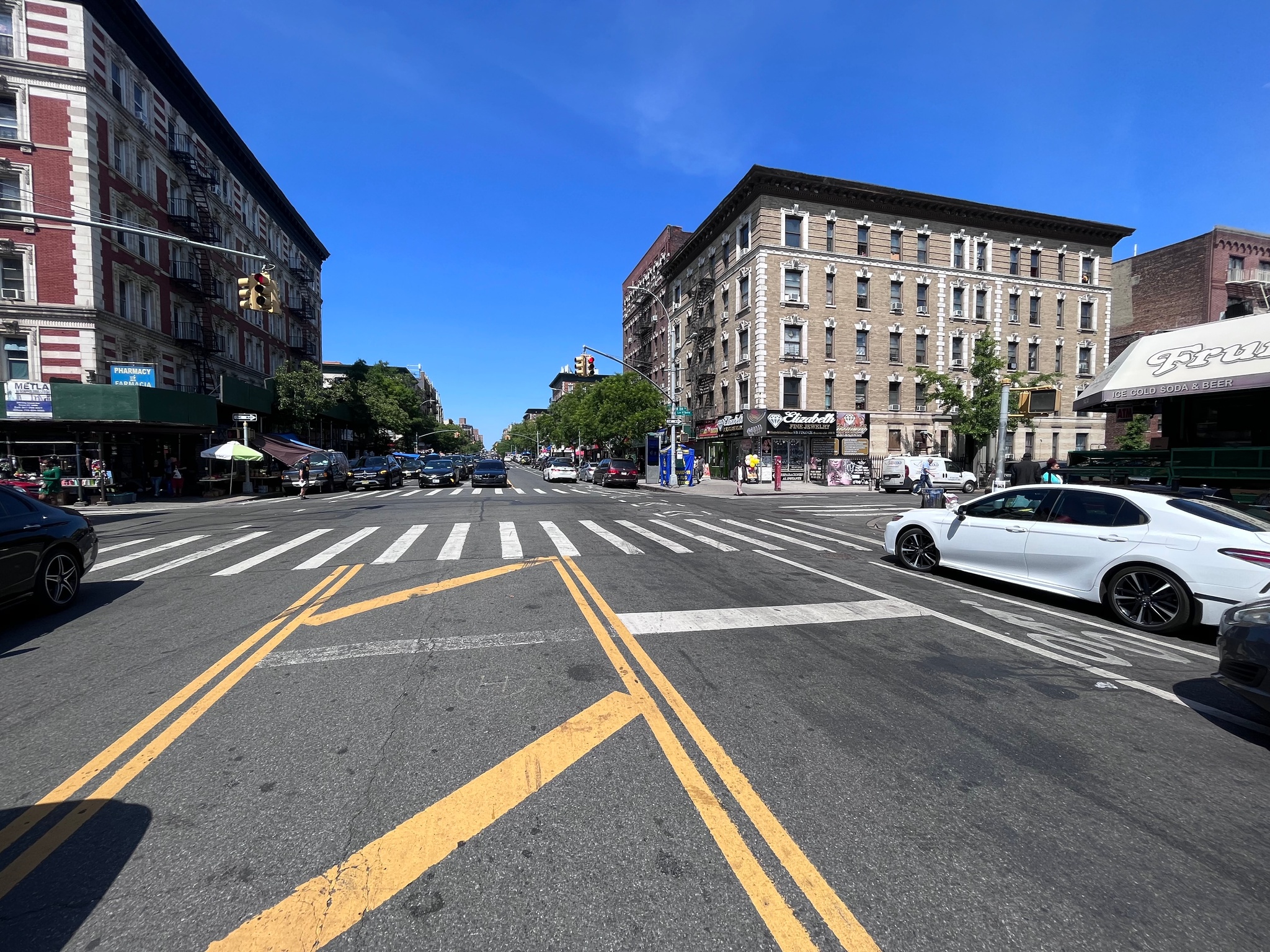 A sweeping view of a city intersection in Washington Heights, Manhattan, with a blue sky above. A white car waits at a traffic light.