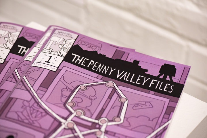 Closeup of a comic book with a purple cover titled "The Penny Valley Files." The cover illustration shows an array of polaroids with tacks and string connecting them in a occult-like arrangement.