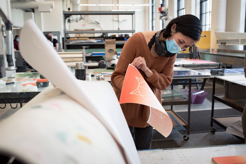 Person pulls an orange and white rainbow rolled print off of a printmaking press table. 