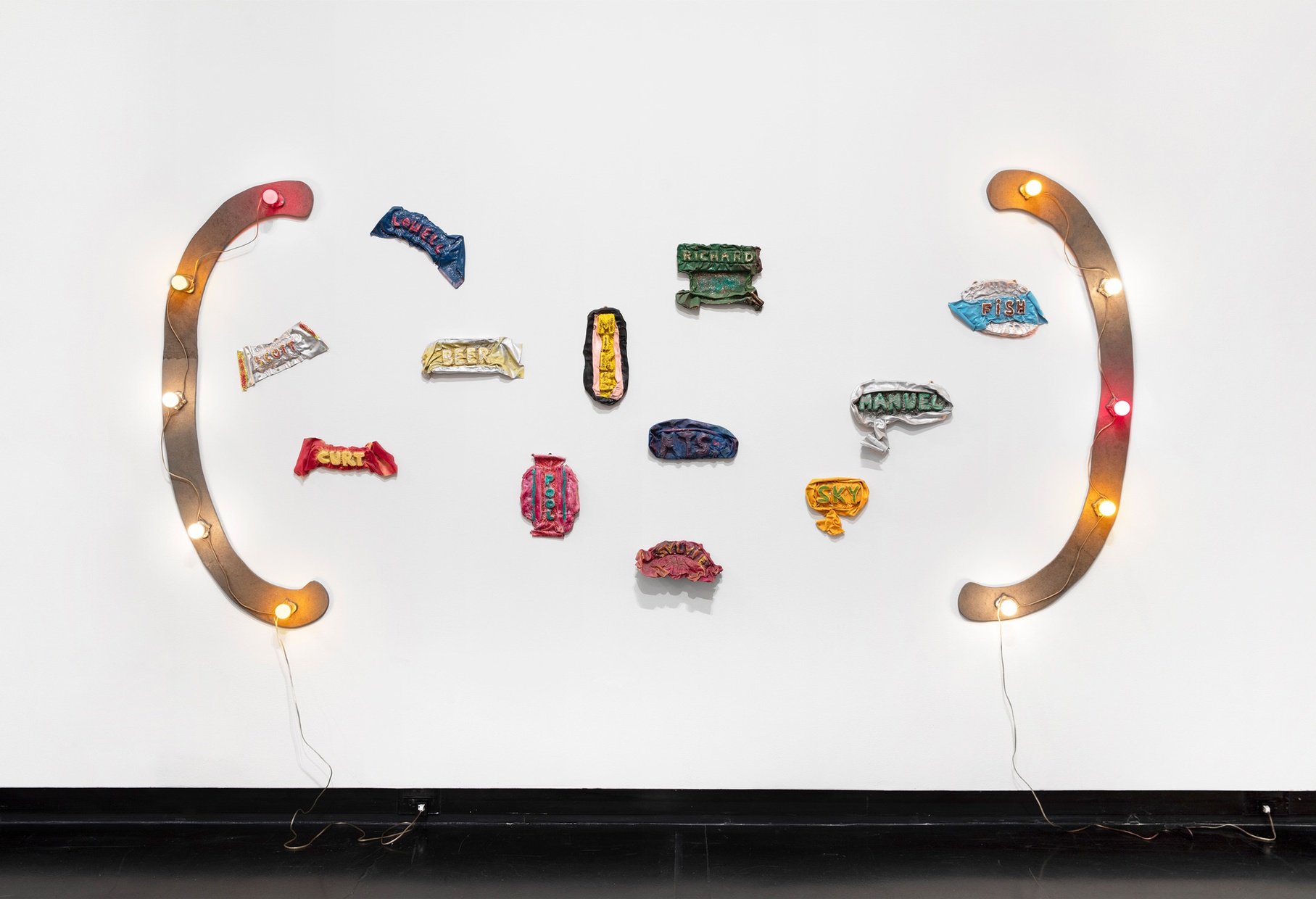 a sculpture, Ree Morton's piece titled "Bozeman, Montana," hangs on a white wall made with enamel, flocking, and glitter on wood and celastic, with bright light bulbs