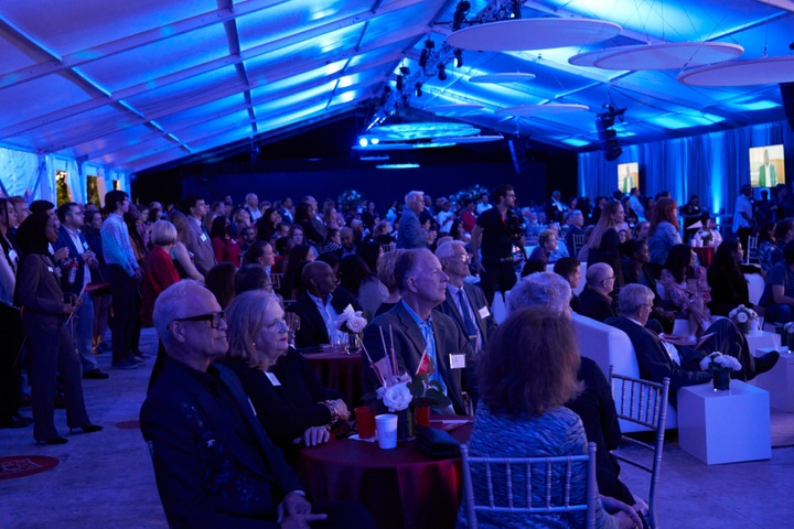 People gathered in a large event tent listening to a speaker. The interior is lit up blue.