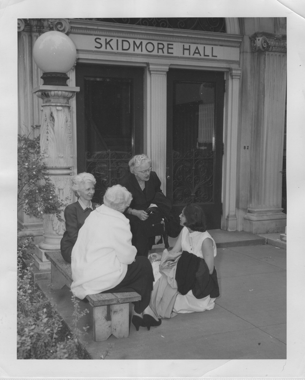 A black and white photograph depicts four light-skinned women in conversation, three of whom are older with white hair and one younger with dark hair, in front of a building with two large doors that says “Skidmore Hall”.
