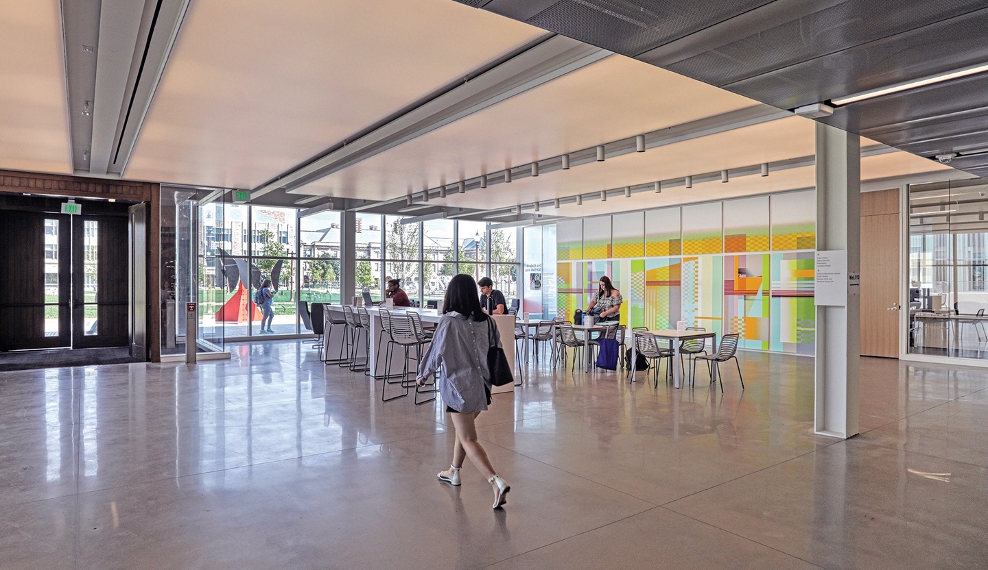 Open commons area filled with light; a bank of floor-to-ceiling windows overlooking green space and an outdoor sculpture sits next to double wooden doors. A colorful mural is on the righthand wall.