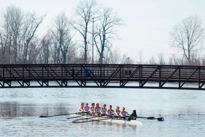 A crew team rows under a bridge on a river in the winter
