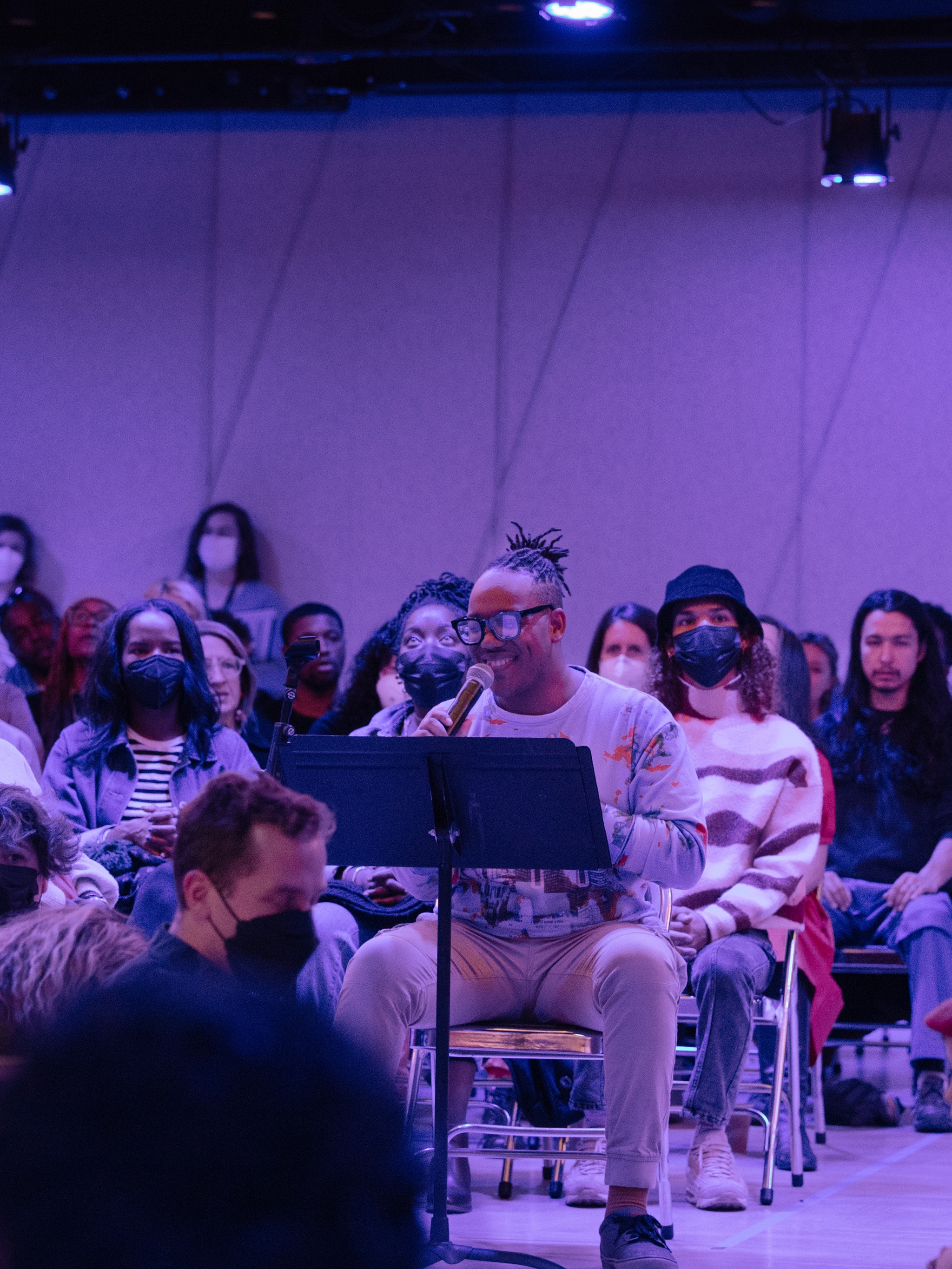A performance space suffused in purple light. Choir members sit in chairs arranged in multiple rows in a circle with audience members mixed in. At the center, Troy Anthony, a Black man and artistic director of The Fire Ensemble, speaks into a microphone.