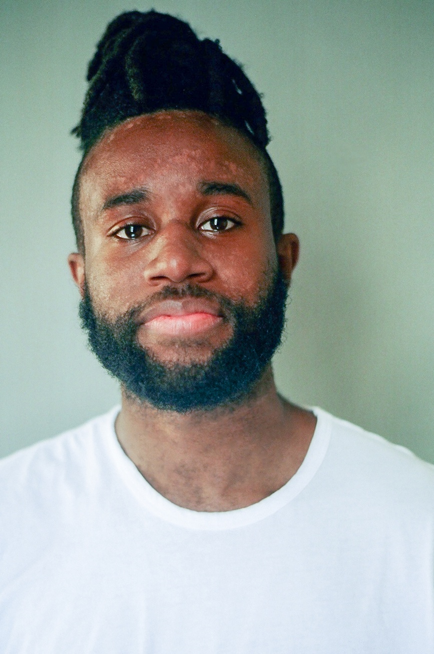 A photo of the artist JJJJJerome Ellis, who has a beard, wears a white t-shirt, and looks into the camera against a pale green background. 