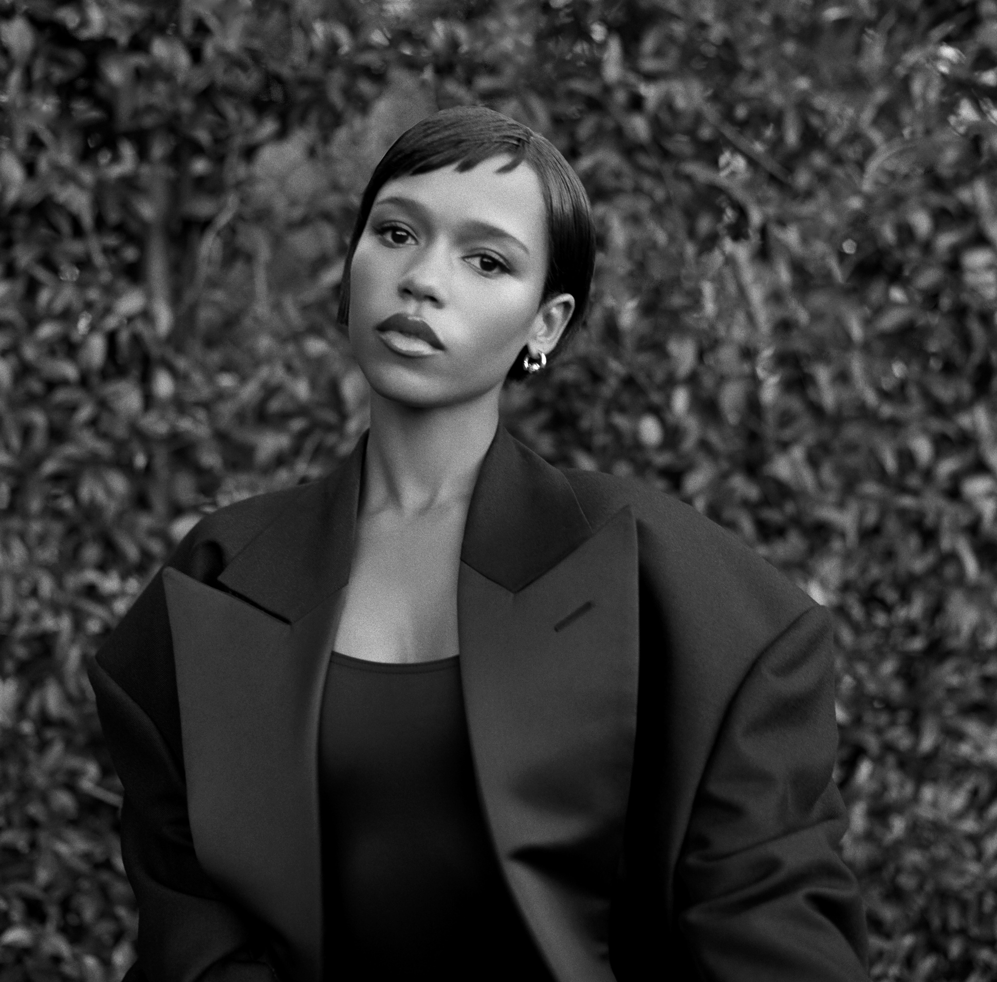 A black and white portrait of actor Taylor Russell. A Black woman, she is seen from the waist up, facing us, against a background of ivy. Taylor has short hair, with bangs swooped to her right across her forehead. She wears a black shoulderless dress with a black dinner jacket with wide shoulders. 
