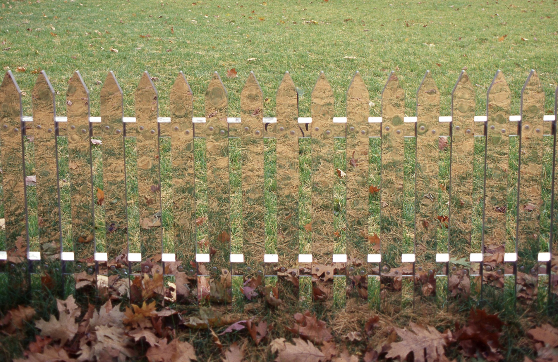 A picket fence made out of mirrors reflects grass and leaves with grass visible behind it.