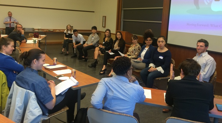 A group of people sit in a row facing another group of people. A presentation screen appears behind them.